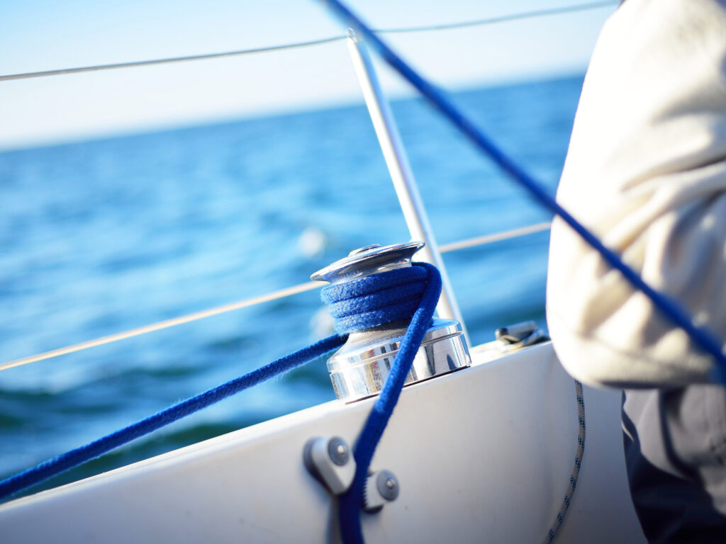 A man sitting next to a staysail winch on a sailboat on a clear day in an open sea